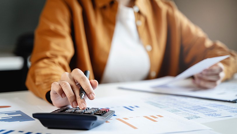 A female accountant types on a calculator while checking on a printed sheet of paper she holds in her hand.
