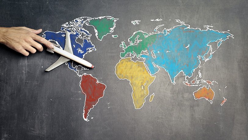 A male adult hand holds a scale model plane over a drawing of a colored world map on a chalkboard, illustrating a
    business overseas expansion.