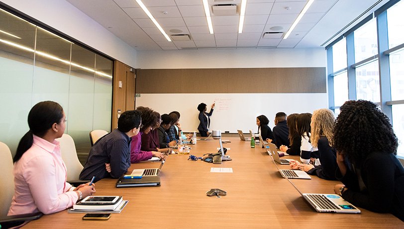 Diverse group of people seated around a large table in a conference room following guidance from a female instructor as
	she points to a whiteboard.