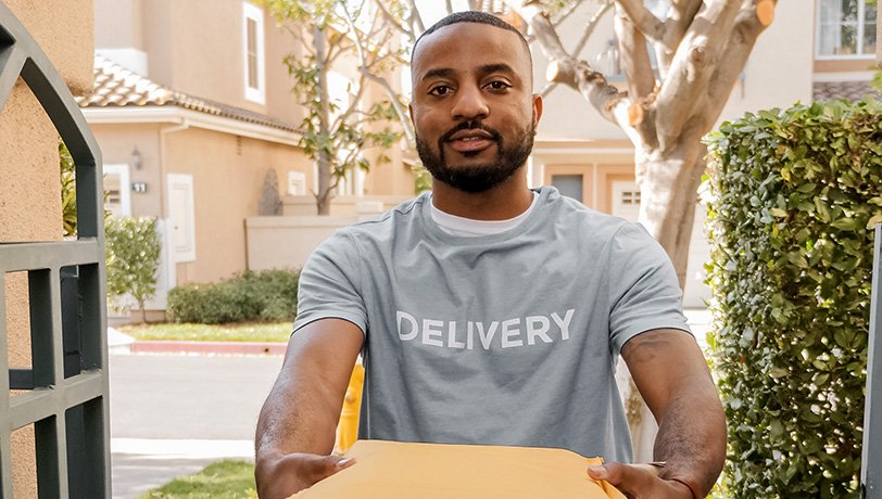 An Afro-American delivery man, wearing a gray t-shirt with the word “Delivery” printed on the chest, delivers a parcel at the front door in a residential street.
