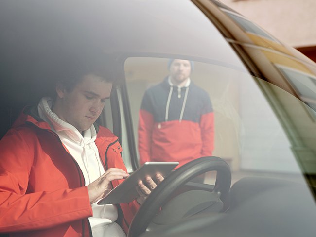 Windshield view of the driver of a small business commercial vehicle, accessing information with a tablet on his hands, while another
		employee appears standing in the background.