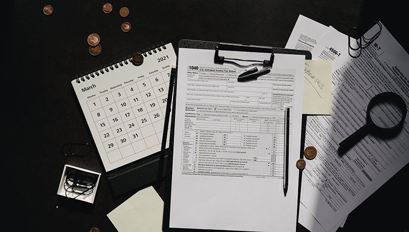 US Tax Forms rest among clipping board, a magnifying glass, a calendar, and coins. Olya Kobruseba - Pexels