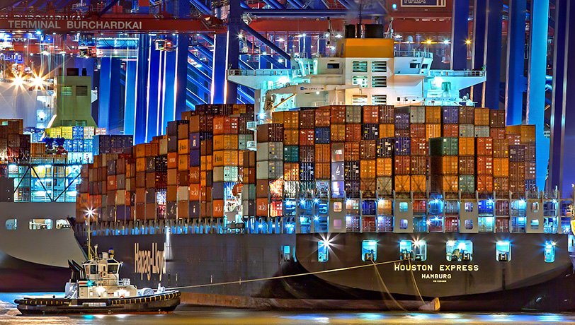 A cargo ship full of containers enters a docking port followed by a tugboat, depicting international trade, most of it led
    by overseas companies.