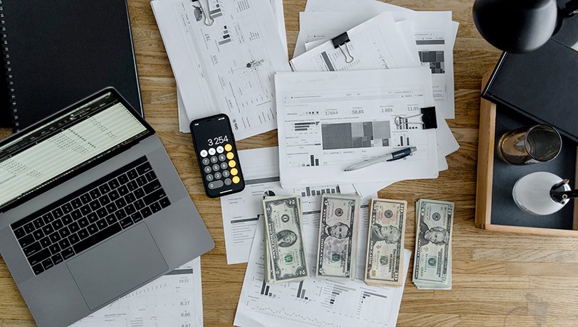 Printed graphs, U. S. dollar bills, the calculator of a mobile phone rest on a desk beside a computer as a reference for payroll software for small business.
